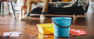 Roof is Leaking due to a Pipe Rupture at Home. Water is dripping into a bucket and a couple is sitting on the couch on the phone with someone for repair.